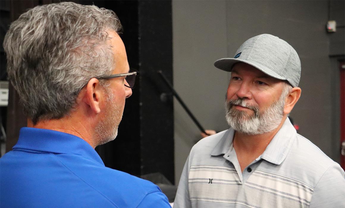 Photo of a person wearing a cap speaking to another person with glasses.